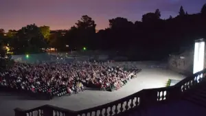Une large foule est assise dans un amphithéâtre en plein air sous un ciel crépusculaire.