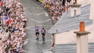 Deux cyclistes roulent sur une route pavée entourés d'une foule enthousiaste portant des chapeaux.