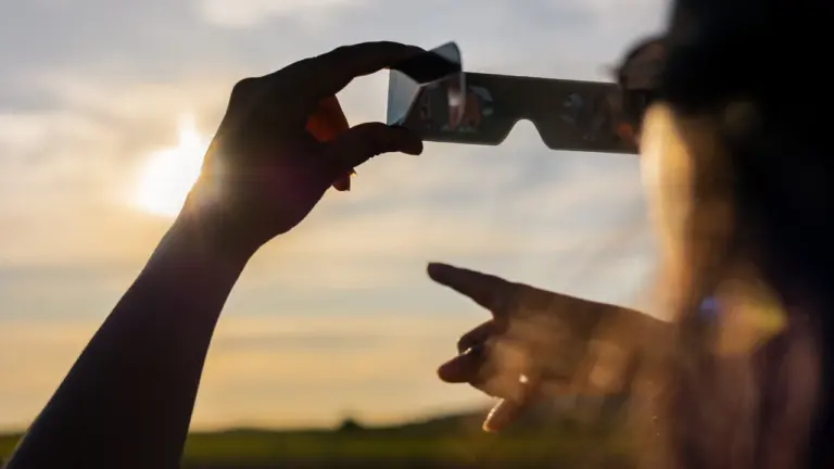 Une personne tient des lunettes de protection solaire vers le soleil couchant à l'horizon.