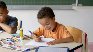 Un jeune garçon souriant écrit sur une feuille dans une salle de classe scolaire.