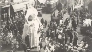 Une foule observe une marionnette géante lors d'une parade dans une rue historique animée.