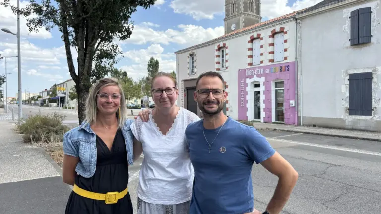 Trois personnes souriantes posent devant un salon de coiffure dans une rue de village.